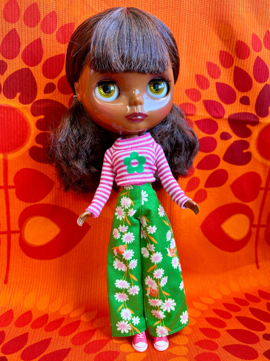 Doll wearing a pink striped shirt and green floral pants against an orange background with red abstract patterns.
