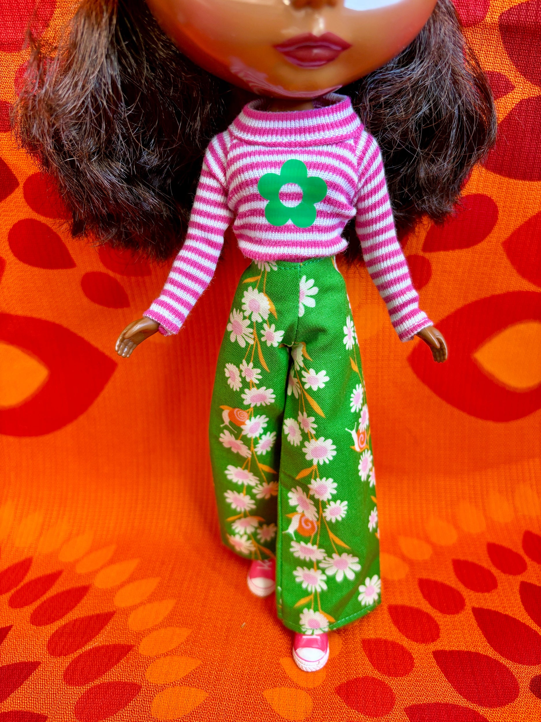 Doll wearing a pink and white striped shirt with a green flower design, and green floral pants on an orange background.