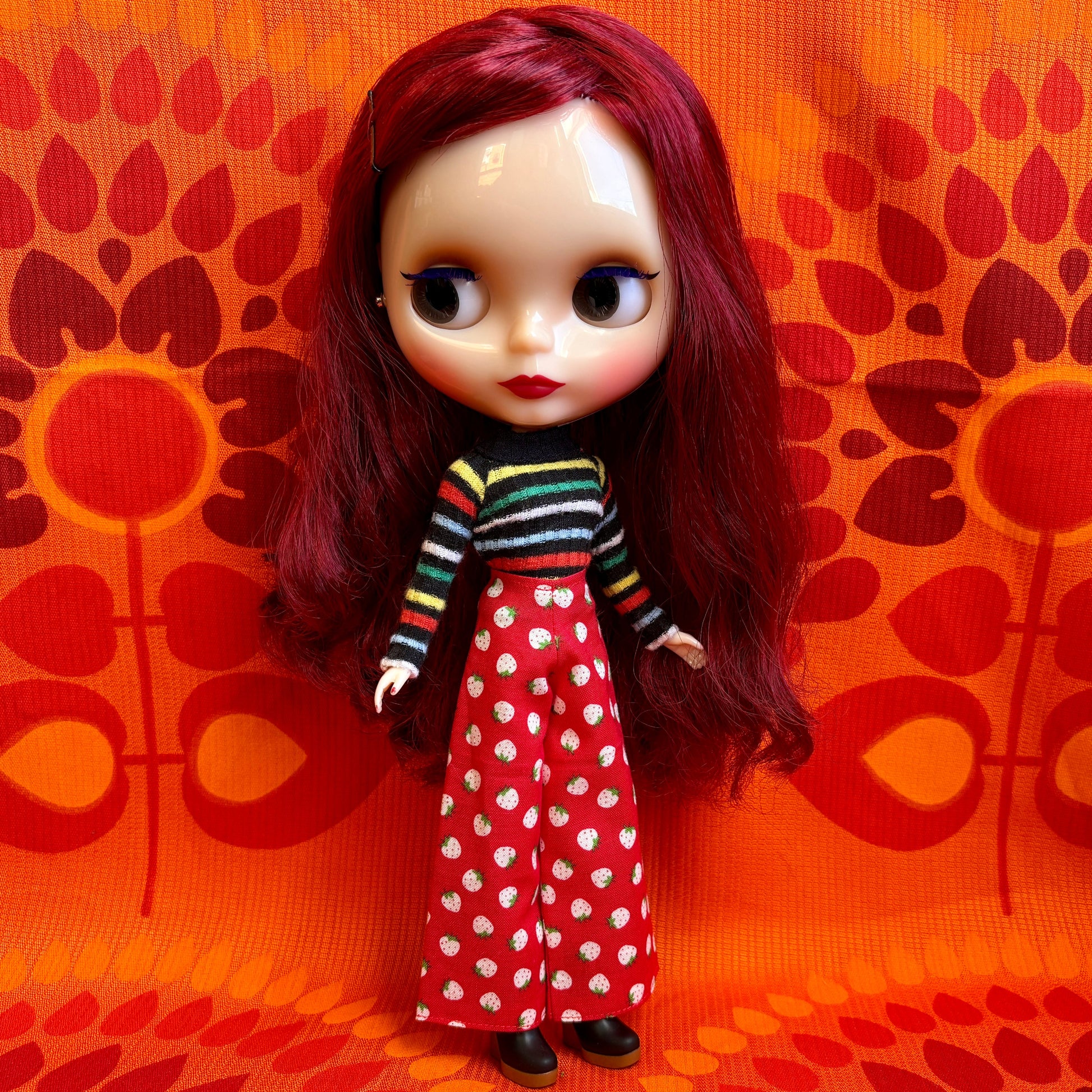 Doll with red hair wearing a striped shirt and polka dot pants against an orange patterned background
