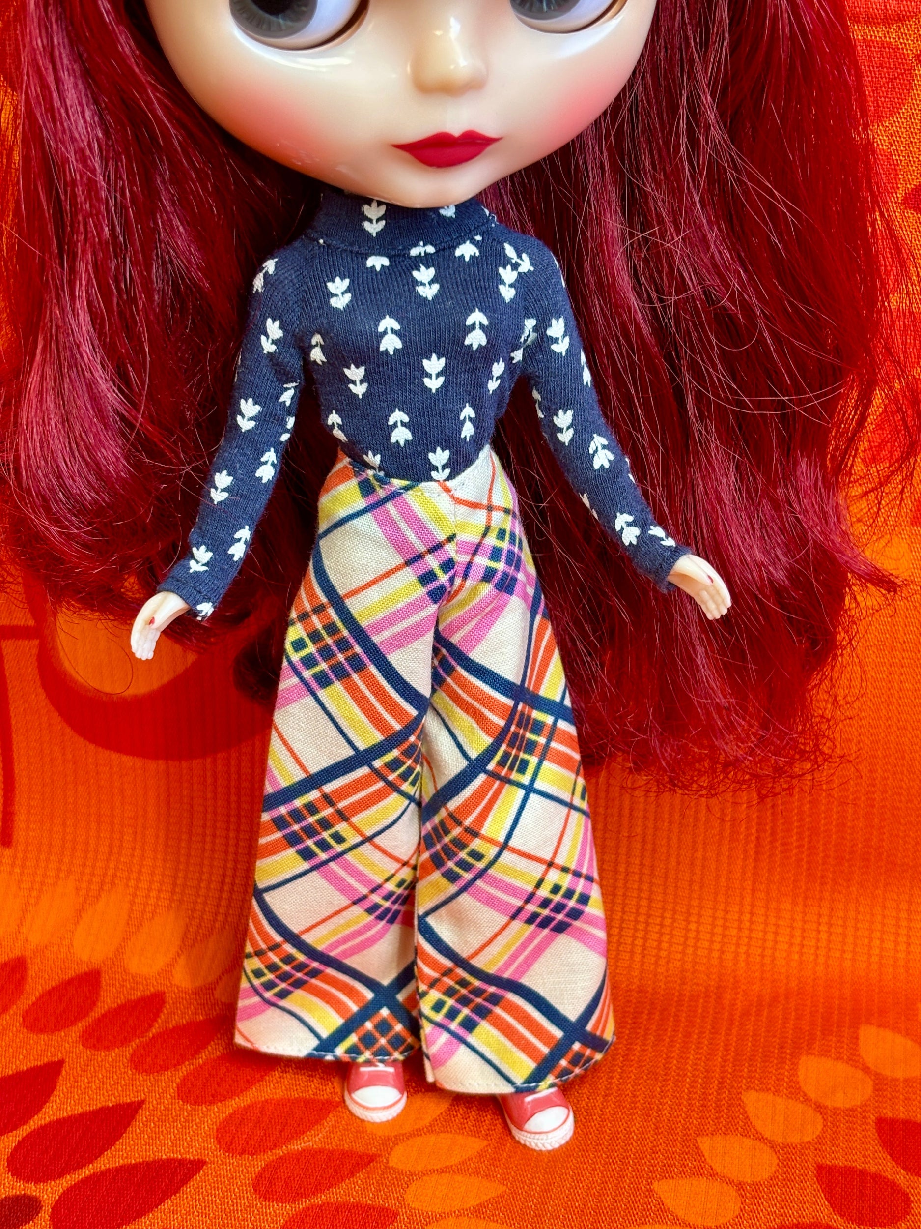 Doll with red hair wearing a navy top with white patterns and colorful plaid pants on an orange background.