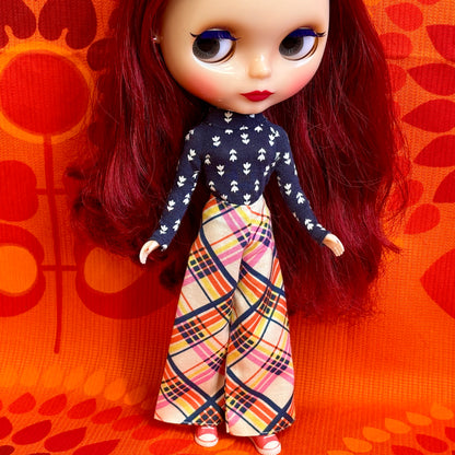 Doll with red hair wearing a navy top and colorful plaid pants against an orange patterned background