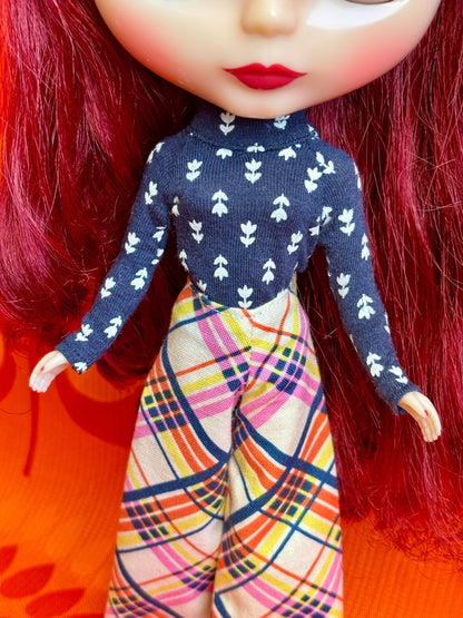 Close-up of a doll wearing a navy blue top with white patterns and colorful plaid pants on an orange background.