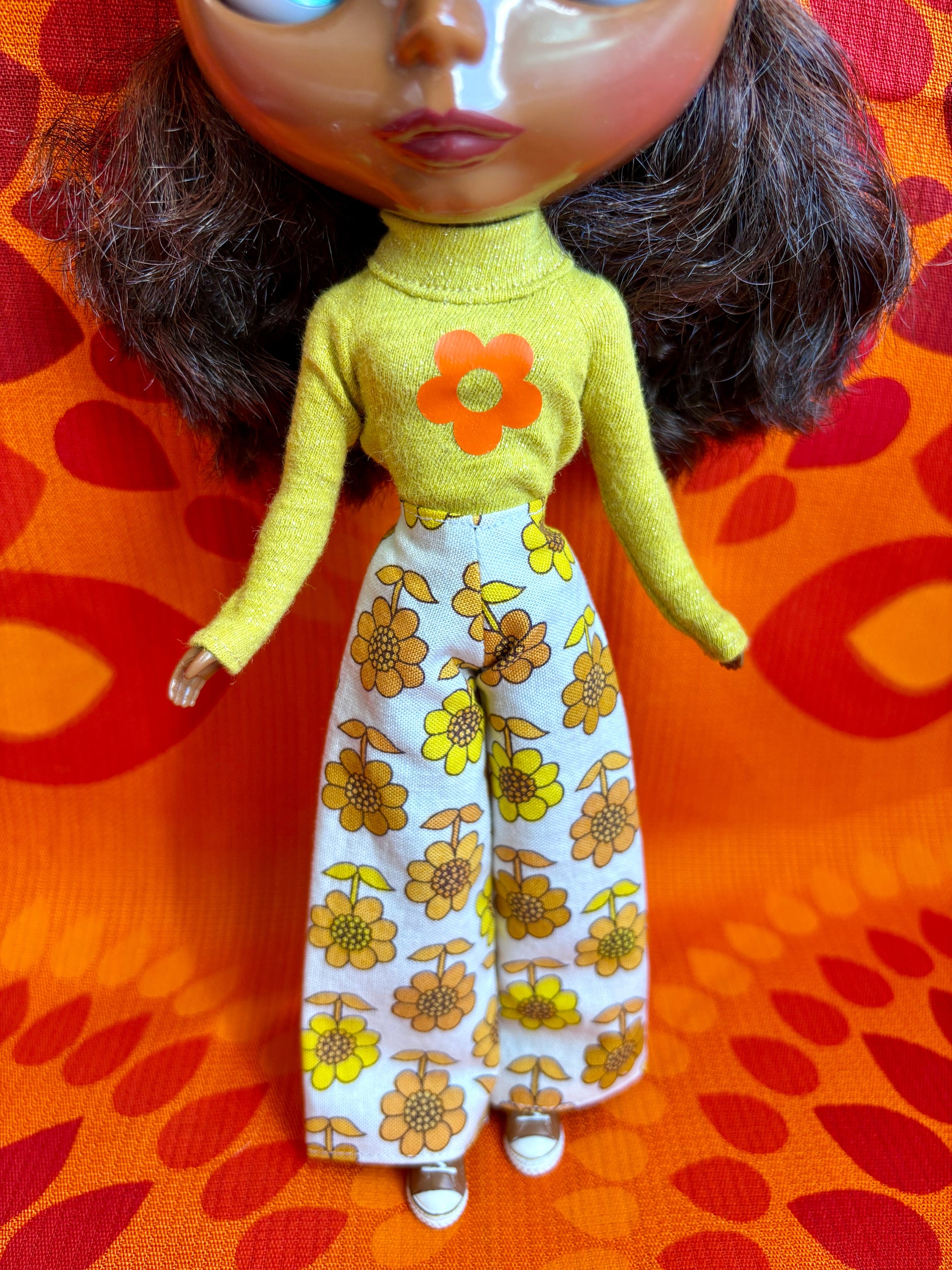 Doll wearing a yellow top with a flower design and floral pants on an orange and red patterned background