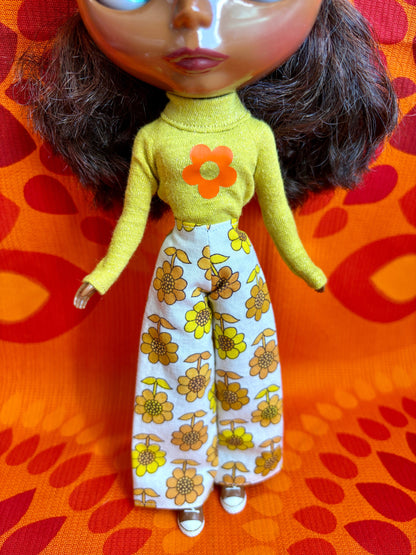 Doll wearing a yellow top with a flower design and floral pants on an orange and red patterned background