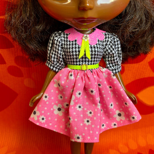 Doll wearing a black and white checkered top with a pink dress featuring floral patterns on an orange background.