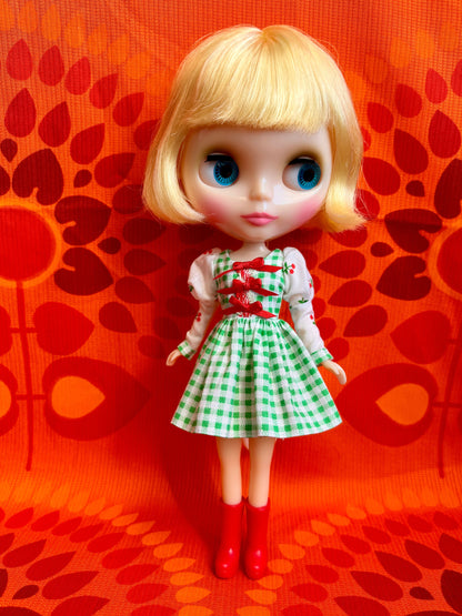 Doll in a green and white checkered dress with red boots on an orange background with red floral patterns.