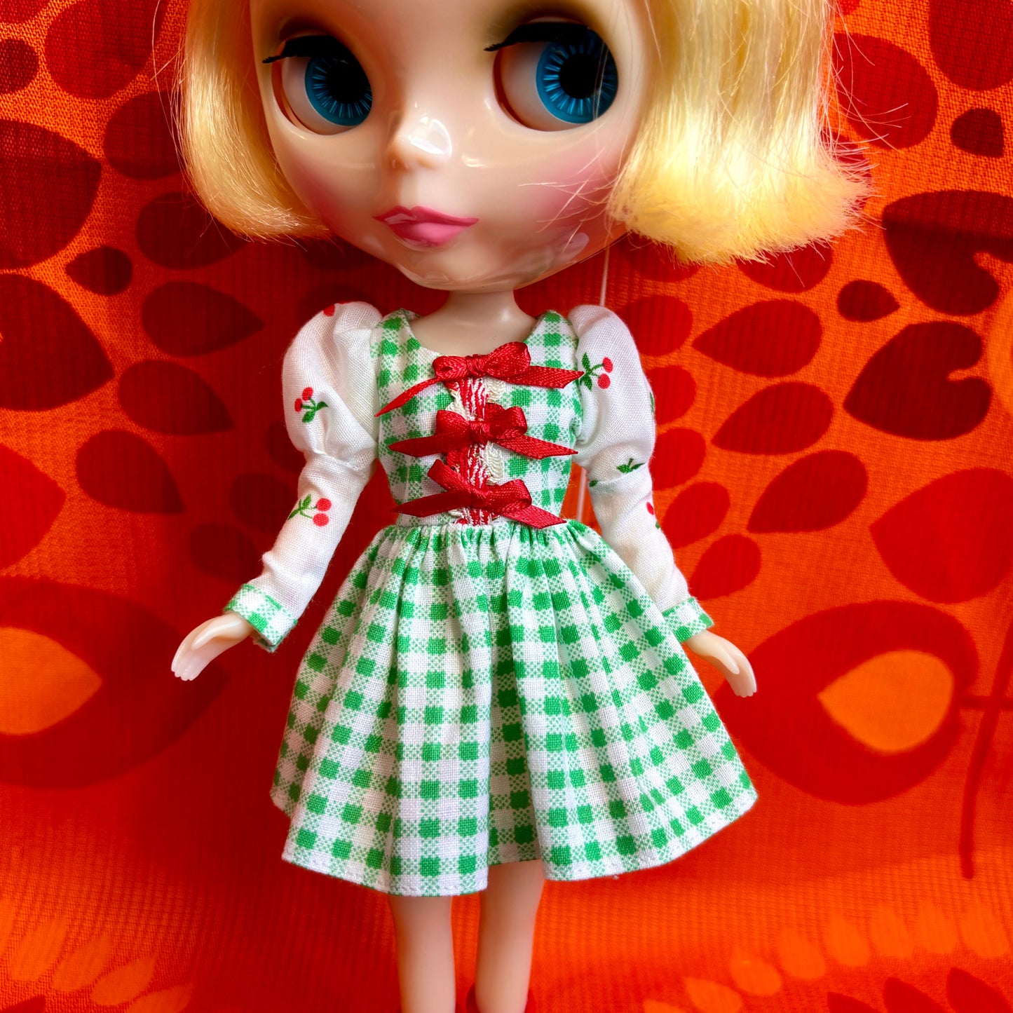 Doll wearing a green and white checkered dress with a red ribbon against an orange background