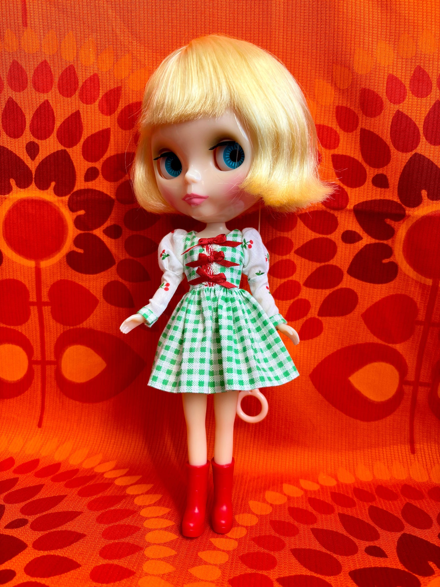  Doll with blonde hair, blue eyes, and a green checkered dress against an orange patterned background.