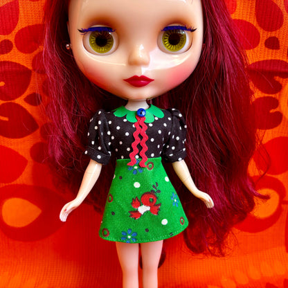 Doll with red hair and a green dress against an orange background with heart patterns