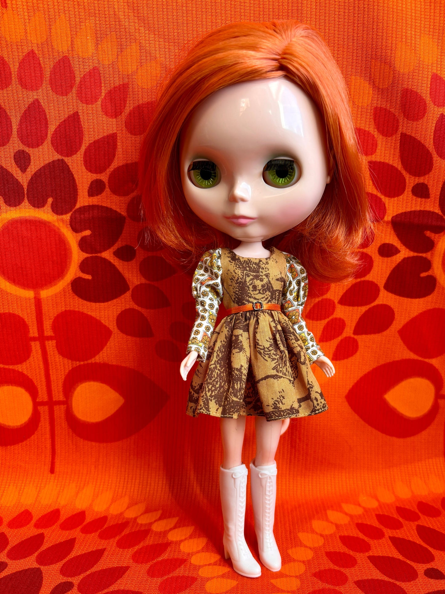 Doll with red hair and a patterned dress standing against an orange background with a leaf design.