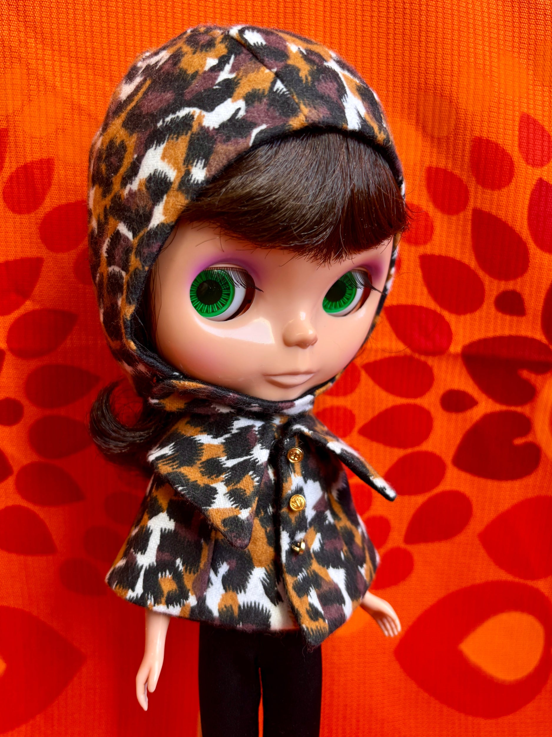 Doll wearing a leopard print helmet against an orange background with red leaf patterns
