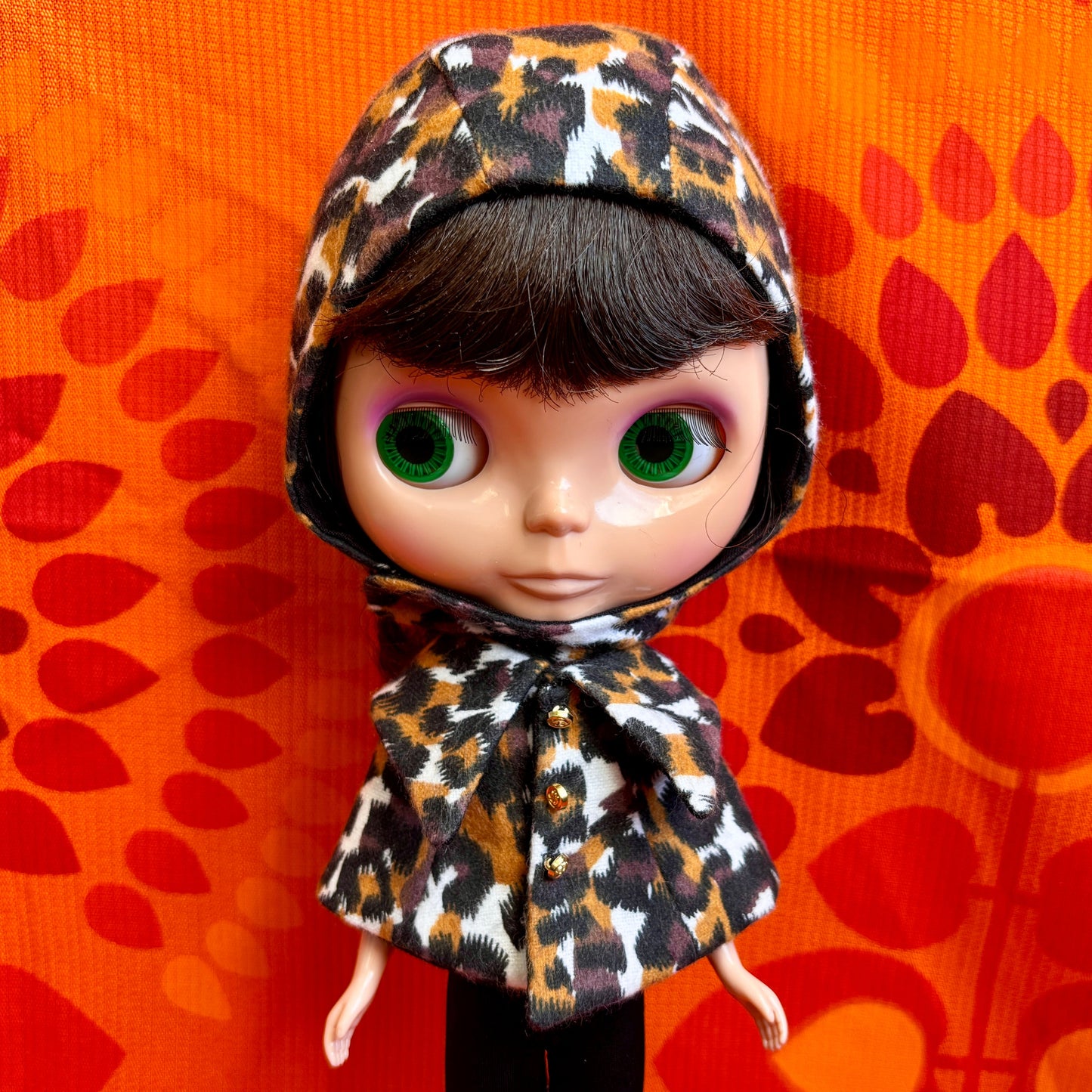 Doll wearing a leopard print hood against an orange background with red leaf patterns