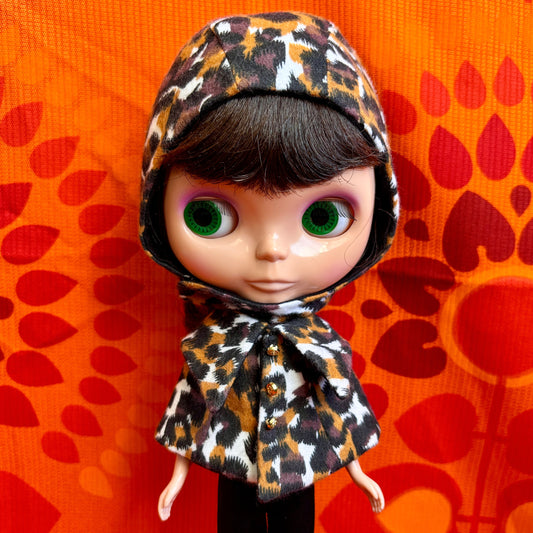 Doll wearing a leopard print hood against an orange background with red leaf patterns