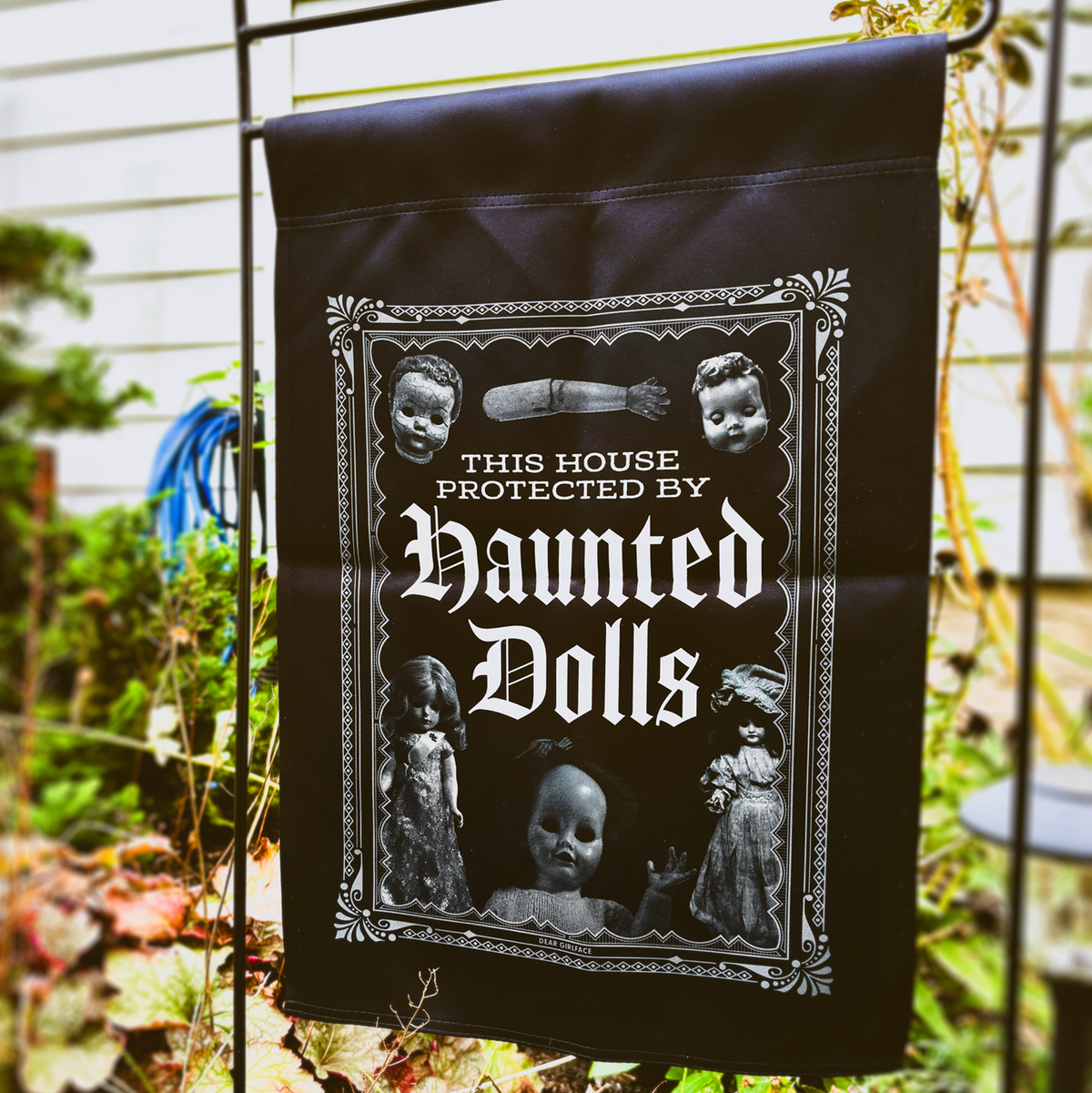A black Halloween garden flag hanging on a black metal fence in a garden. The flag features a gothic white graphic with the text 'THIS HOUSE PROTECTED BY Haunted Dolls' and illustrations of vintage dolls and a creepy doll face.
